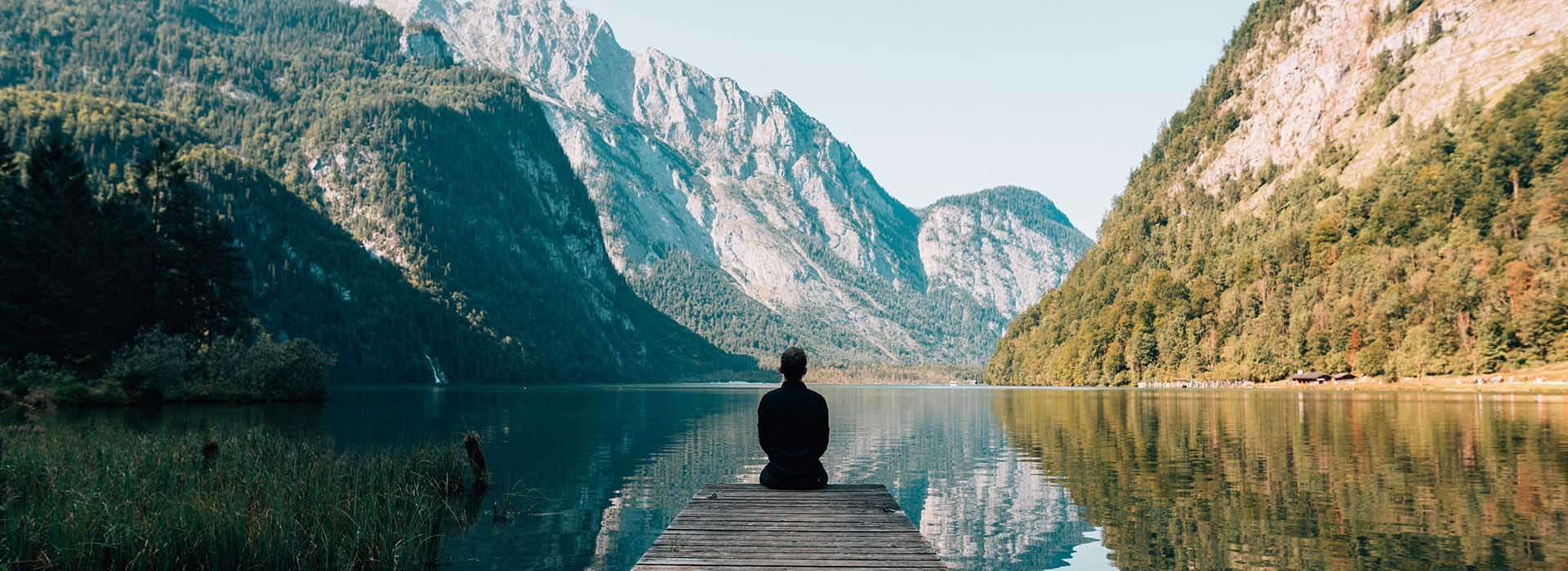person sitting at the end of a pier in a serene mountain setting
