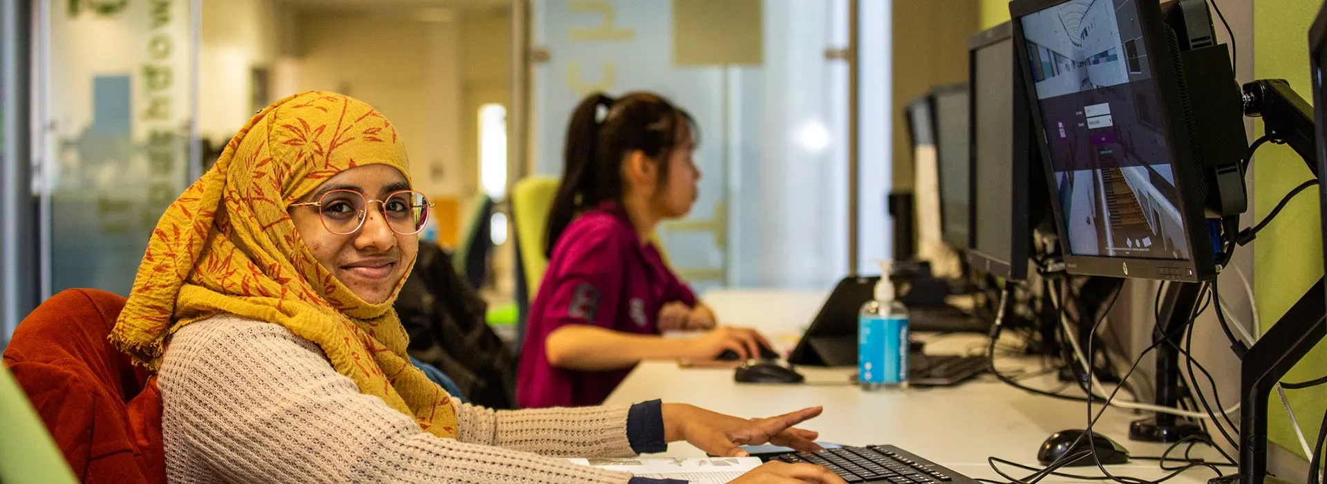 student wearing a headscarf sitting at a computer desk smiling