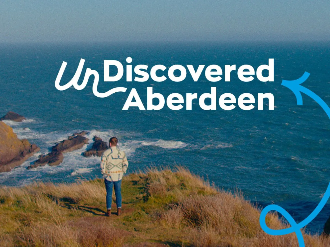 A man stands looking out from Dunnottar Castle, with UnDiscovered Aberdeen branding in the foreground