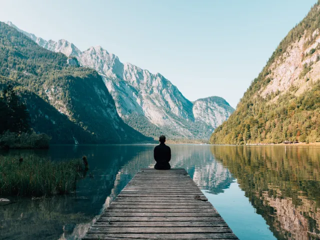 person sitting at the end of a pier in a serene mountain setting