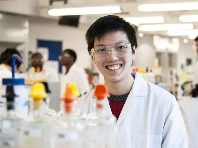 student in a lab coat and safety glasses smiling in front of science equipment