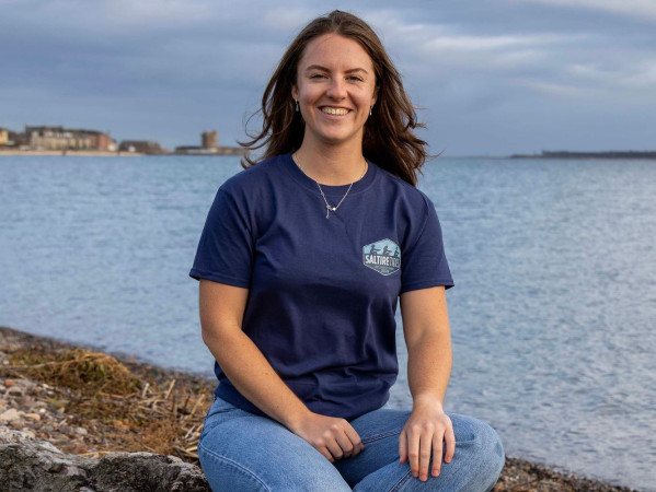 A woman wearing a dark blue t-shirt sitting on a rock next to water