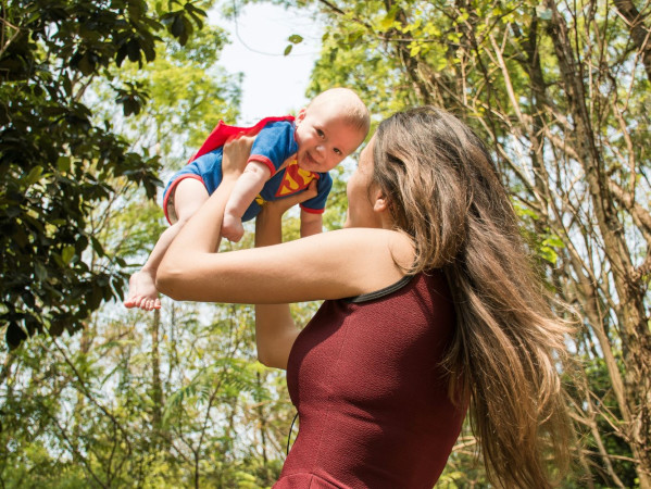 Image shows mother lifting a baby in the air, wearing a superman baby grow - Credit: Valeria Zoncoll on Unsplash