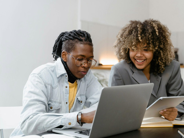 Two people talk while looking at a laptop and tablet